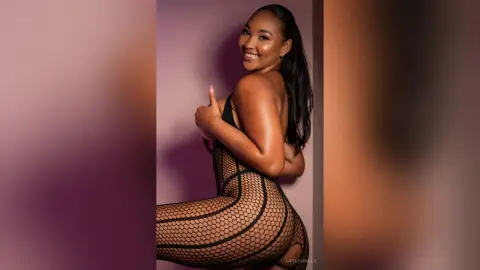 Smiling Black woman in a black fishnet bodysuit posing confidently against a soft purple background.