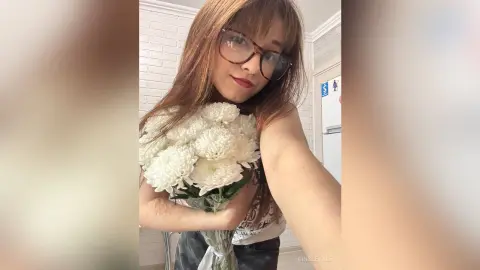 Smiling young woman with glasses holding a large bouquet of white chrysanthemums in a bright indoor setting.