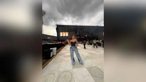 Red-haired woman in a black corset and baggy jeans standing at an outdoor music festival under a cloudy sky.