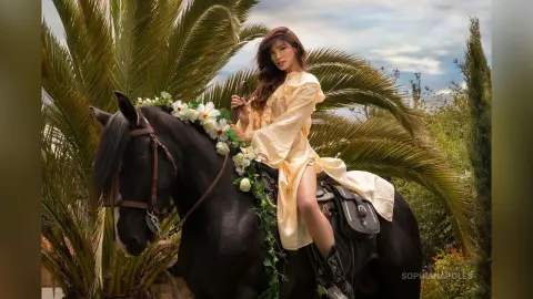 SophiaNapoles sits atop a black horse wearing a light yellow dress, posing outdoors against a backdrop of large palm leaves.