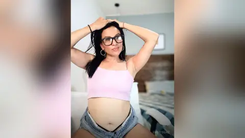 Smiling woman in pink crop top and denim shorts posing in a bright bedroom.