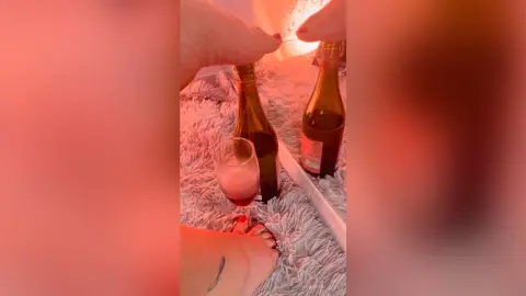 Two feet relaxing on a white shag rug next to beer bottles under warm red ambient lighting.