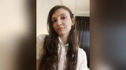 MillieStrife poses with long, wavy brown hair wearing a white button-up shirt against a neutral wall and dark curtain background.
