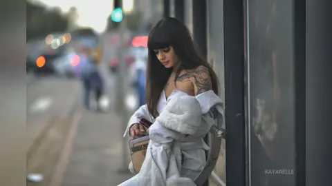 Stylish brunette woman with shoulder tattoos in a white fuzzy coat sitting on a city street bench.