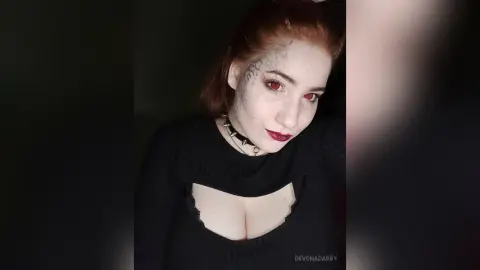 Gothic woman with red eyes and facial tattoos wearing a black choker in a moody, dark setting.