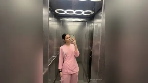 Woman in pink medical scrubs taking a mirror selfie in a modern elevator.