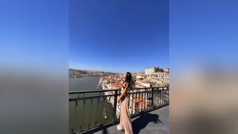 LucyMartin stands on a balcony overlooking a river and city, wearing a black bra and light tan trousers.