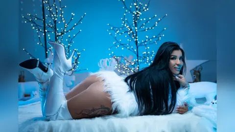 MissyJolie lies on a white furry surface wearing white thigh-high boots and white furry lingerie, surrounded by lit decorative trees.