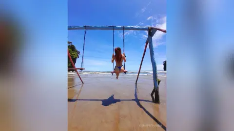 Woman in a blue bikini sitting on a wooden swing at a tropical beach under a clear blue sky.