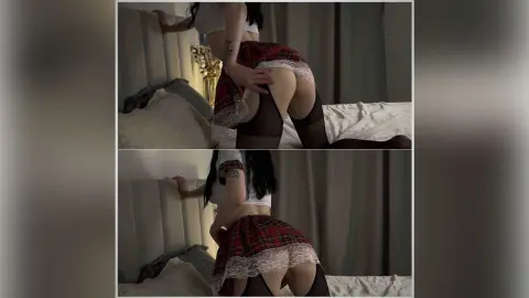 Woman in a red plaid skirt and white top posing on a bed in a moody, dimly lit bedroom setting.