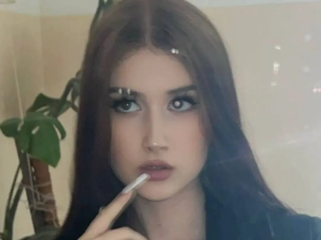 Young woman with long brown hair and soft makeup looking thoughtfully in a mirror near indoor plants.