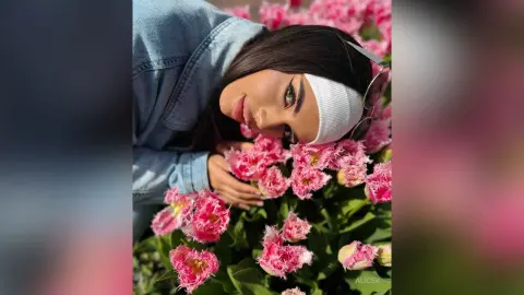 Young woman in denim jacket relaxing amidst vibrant pink flowers in a sunny garden setting.