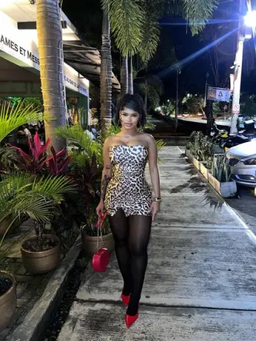 Woman in leopard print mini dress and black tights walking outdoors at night near tropical plants.