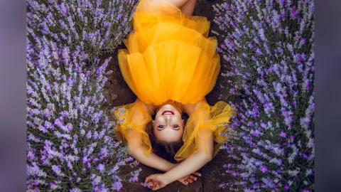 AmelieSkylar lies on the ground wearing a bright yellow tulle dress, surrounded by purple lavender flowers.