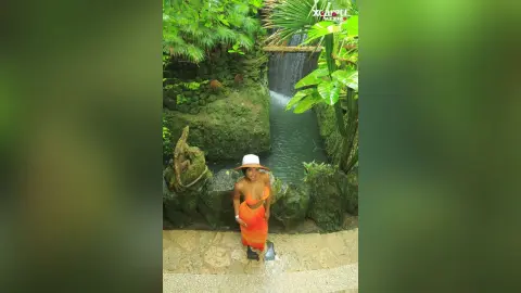 NohomiCambel stands in front of a small waterfall wearing an orange halter-style top and matching skirt or wrap.