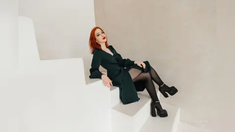 Elegant redhead woman in a dark green dress sitting on white stairs in a minimalist modern setting.