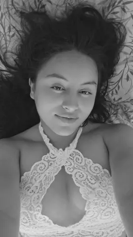 Smiling young woman in a white lace halter top lying on a floral patterned bed in a serene black and white portrait.