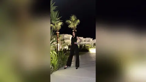 Stylish woman in a black jumpsuit standing by palm trees at a luxury resort at night.