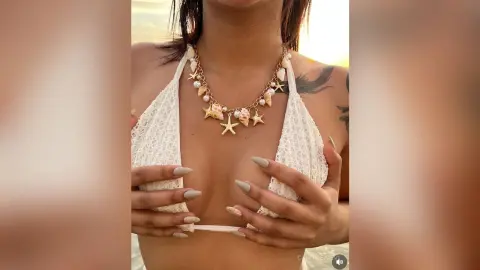Woman in a white lace bikini wearing a seashell necklace at a sunny beach setting.