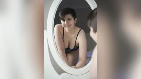 Smiling young woman in a black lace bra looking at her reflection in a round white mirror in a bright room.