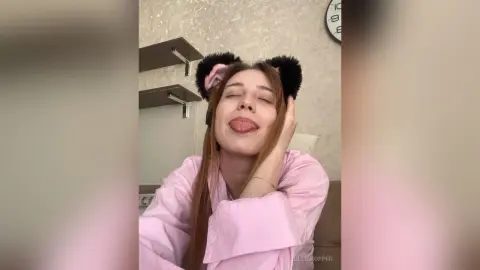 Cheerful young woman in pink pajamas wearing black fuzzy headband relaxing in a cozy bedroom setting.