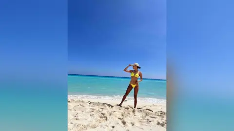 Confident woman in a yellow bikini posing on a sunny tropical beach with turquoise ocean water.
