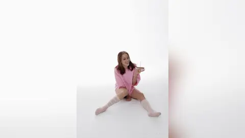 NoellaGlowinski sits on a white floor wearing a pink shirt and light pink socks, holding a small object in her hands.