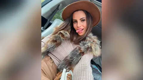 Smiling woman in a beige sweater and brown hat with a faux fur collar sitting in a car.