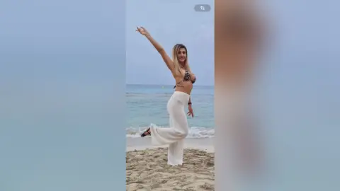 Cheerful woman in a black bikini top and white wide-leg pants posing on a sunny beach by the ocean.
