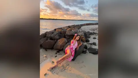 Young woman in a pink summer dress relaxing on a sandy beach by large rocks at sunset.