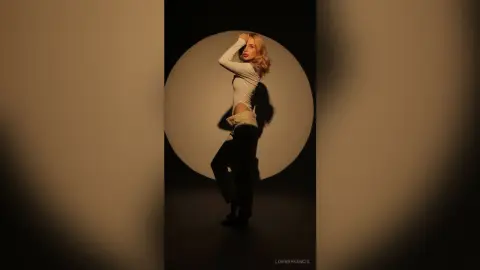 Blonde woman in a white long-sleeve crop top and black trousers posing against a circular spotlight backdrop.