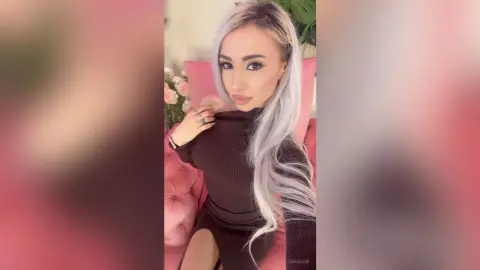 Stylish woman with long silver hair wearing a brown ribbed dress sitting on a pink velvet sofa.