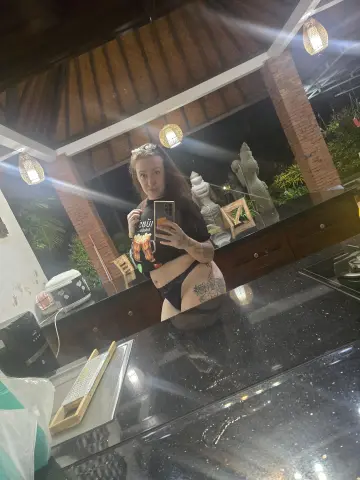Woman in black t-shirt and bikini bottoms taking a mirror selfie in a tropical outdoor kitchen setting.
