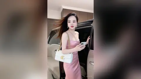 EnyaLin stands next to a car wearing a pink strapless dress and holding a white purse and phone.