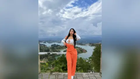 Stylish woman in orange trousers and white crop top posing against a scenic cloudy mountain lake view.