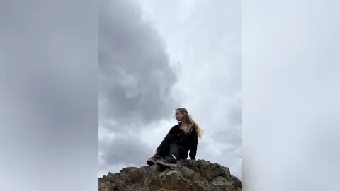 Blonde woman in black outfit sitting on rocks under a moody, cloudy sky.