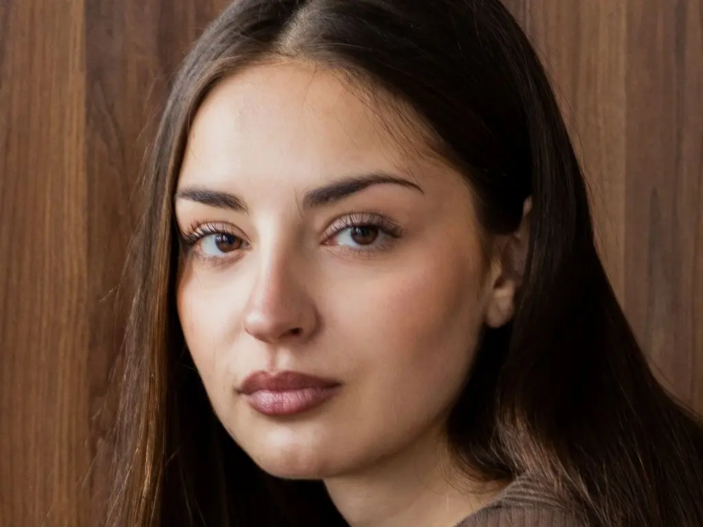 Close-up portrait of a young woman with long brown hair looking serene against a warm wooden background.