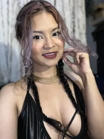 Hathorlove smiles while wearing a black leather halter top and gold choker, showing off her long, wavy purple-tinted hair and dark braids.