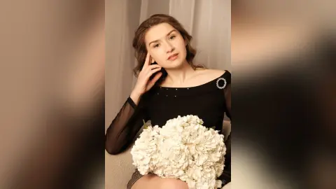 Elegant woman in a black long-sleeve dress holding a white flower bouquet in a soft, dreamy indoor setting.
