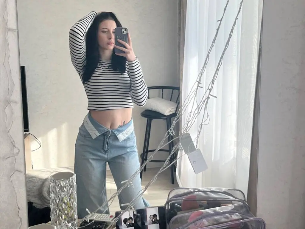 Young woman in a striped long-sleeve crop top and light wash denim jeans taking a mirror selfie in a bright room.