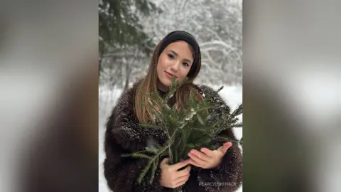 Young woman in a black faux fur coat holding evergreen branches in a snowy winter forest setting.