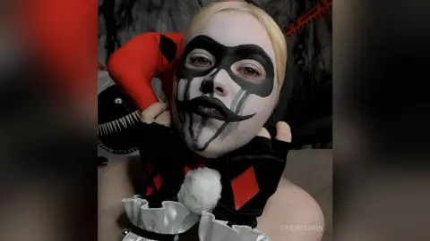 Woman in Harley Quinn cosplay with dramatic face paint and jester outfit in a dark moody setting.