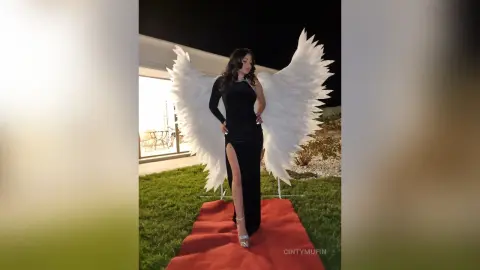 Woman in elegant black evening gown with large white angel wings standing on a red carpet at night.