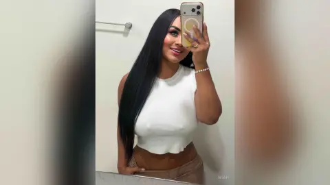 Smiling woman with long black hair in a white sleeveless crop top taking a mirror selfie in a bright bathroom.