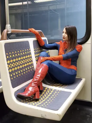 JesseSpark wears a blue and red superhero bodysuit and red boots while sitting on a bus seat.