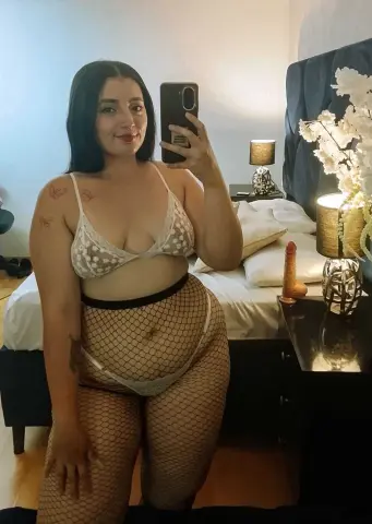 Woman in white lace bra and fishnet tights posing in a cozy bedroom setting.