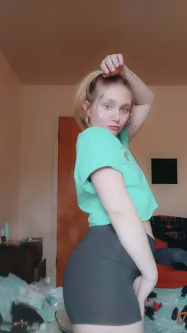 SummerBrookes poses in a mint green t-shirt and tight grey shorts, running her hand through her blonde hair in a bedroom setting.