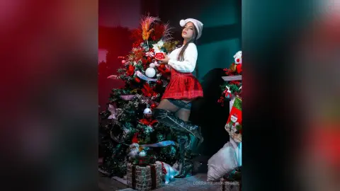Young woman in festive red skirt and white sweater posing by a decorated Christmas tree in a cozy holiday mood.