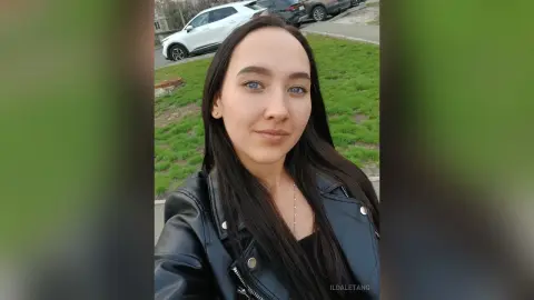 Smiling young woman with long dark hair wearing a black leather jacket in an outdoor park setting.