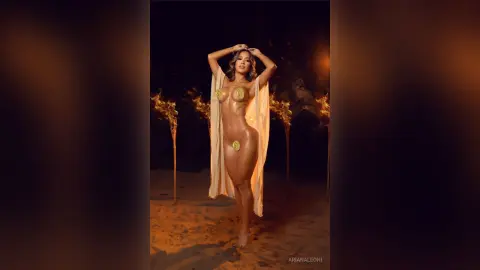 Glamorous woman in a gold bikini and flowing white robe posing amidst glowing torches at night.